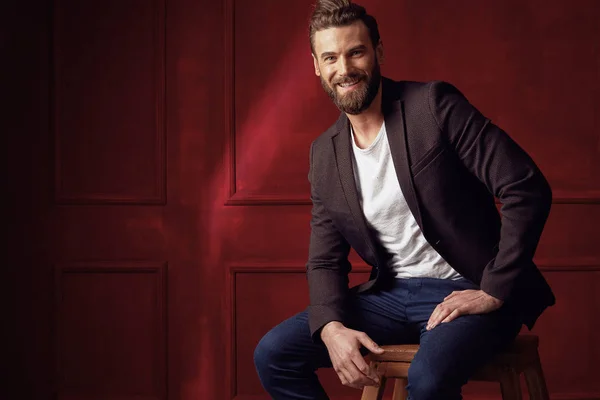 Attractive young beautiful and confident bearded man, wearing white shirt and brown jacket, sitting on wooden chair in studio with dark red background, and smiling — Stock Photo, Image