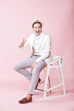 Beyaz ceket, beyaz gömlek, gri pantolon ve açık pembe renkli Kişilik koltuğa oturan beyaz şapka giyen genç gülümseyen adam