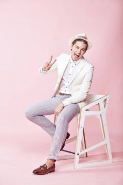 Beyaz ceket, beyaz gömlek, gri pantolon ve açık pembe renkli Kişilik koltuğa oturan beyaz şapka giyen genç çekici gülümseyen adam