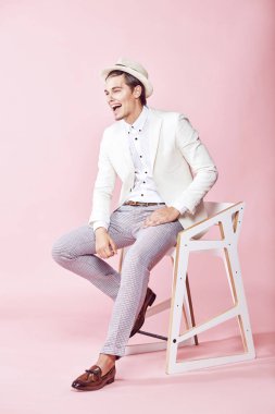 Genç çekici mutlu gülümseyen adam beyaz ceket, beyaz gömlek, gri pantolon ve beyaz şapka ile açık pembe renkli stüdyo sandalyede oturan giymiş