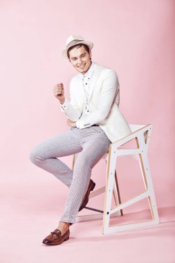 Genç güzel gülümseyen erkek model beyaz ceket, beyaz gömlek, gri pantolon ve beyaz şapka ile açık pembe renkli stüdyo sandalyede oturan giymiş
