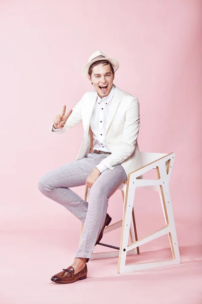 Pemuda tersenyum mengenakan jaket putih, kemeja putih, celana abu-abu dan topi putih duduk di kursi di studio dengan latar belakang merah muda yang ringan — Stok Foto