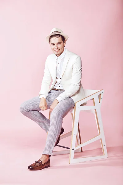 Unga attraktiva leende man klädd i vit kavaj, vit skjorta, grå byxor och vit hatt sitter på stolen i studio med ljusrosa bakgrund — Stockfoto