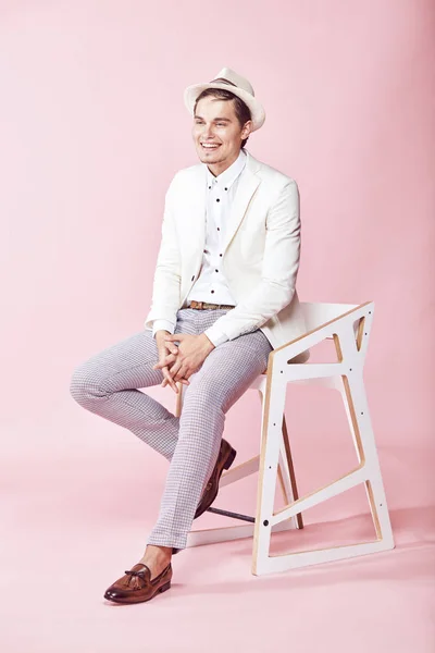 Unga vackra glada leende man klädd i vit kavaj, vit skjorta, grå byxor och vit hatt sitter på stolen i studio med ljusrosa bakgrund — Stockfoto