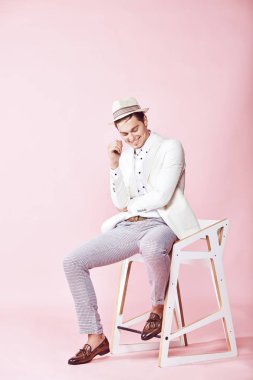 Beyaz ceket, beyaz gömlek, gri pantolon ve açık pembe renkli Kişilik koltuğa oturan beyaz şapka giyen genç güzel mutlu modern erkek modeli