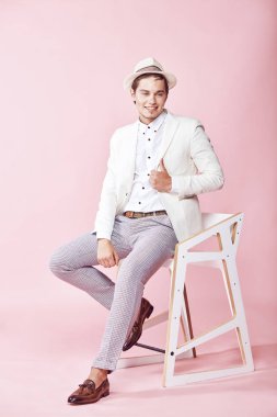 Genç güzel gülümseyen erkek model beyaz ceket, beyaz gömlek, gri pantolon ve beyaz şapka ile açık pembe renkli stüdyo sandalyede oturan giymiş
