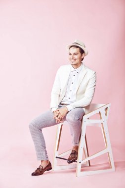 Beyaz ceket, beyaz gömlek, gri pantolon ve açık pembe renkli Kişilik koltuğa oturan beyaz şapka giyen genç güzel mutlu modern erkek modeli