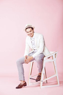 Genç yakışıklı mutlu modern adam beyaz ceket, beyaz gömlek, gri pantolon ve beyaz şapka ile açık pembe renkli stüdyo sandalyede oturan giymiş