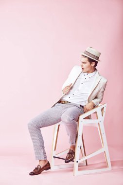 Beyaz ceket, beyaz gömlek, gri pantolon ve açık pembe renkli Kişilik koltuğa oturan beyaz şapka giyen genç yakışıklı mutlu modern erkek modeli