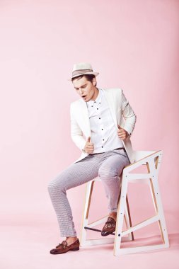 Beyaz ceket, beyaz gömlek, gri pantolon ve açık pembe renkli Kişilik koltuğa oturan beyaz şapka giyen genç yakışıklı mutlu modern erkek modeli