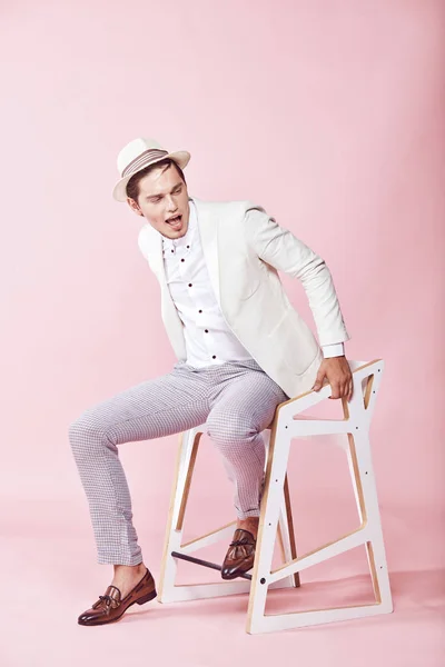 Muda cantik bahagia model pria modern mengenakan jaket putih, kemeja putih, celana abu-abu dan topi putih duduk di kursi di studio dengan latar belakang merah muda yang ringan — Stok Foto
