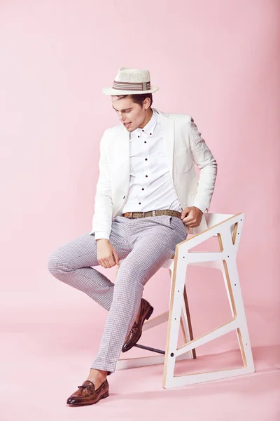 Unga vackra leende manlig modell bär vit jacka, vit skjorta, grå byxor och vit hatt sitter på stolen i studio med ljusrosa bakgrund — Stockfoto