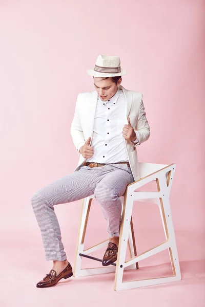 Unga vackra moderna manlig modell bär vit jacka, vit skjorta, grå byxor och vit hatt sitter på stolen i studio med ljusrosa bakgrund — Stockfoto