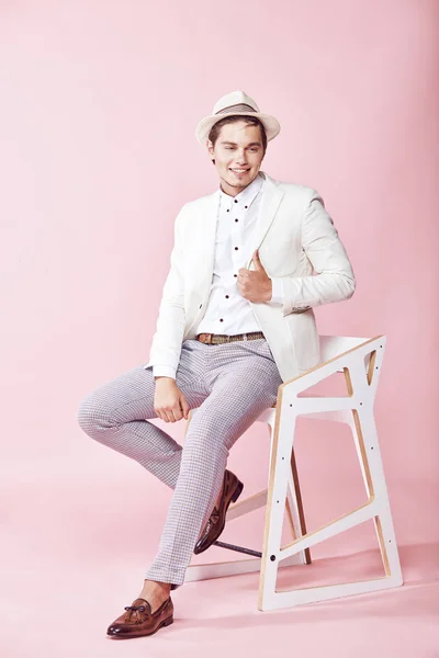 Muda cantik tersenyum model laki-laki mengenakan jaket putih, kemeja putih, celana abu-abu dan topi putih duduk di kursi di studio dengan latar belakang merah muda yang ringan — Stok Foto