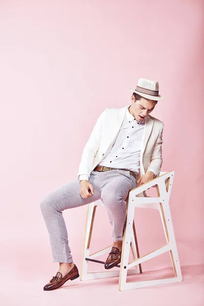 Unga stiliga glad moderna manlig modell bär vit jacka, vit skjorta, grå byxor och vit hatt sitter på stolen i studio med ljusrosa bakgrund — Stockfoto