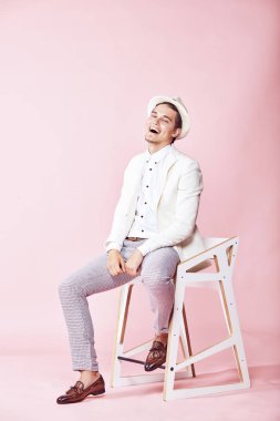 Beyaz ceket, beyaz gömlek, gri pantolon ve açık pembe renkli Kişilik koltuğa oturan beyaz şapka giyen genç yakışıklı mutlu gülen modern insan