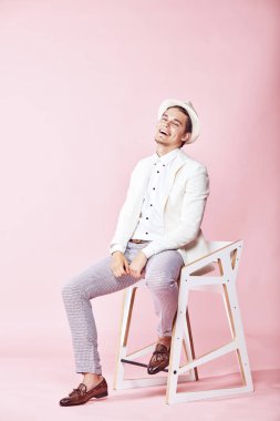 Beyaz ceket, beyaz gömlek, gri pantolon ve açık pembe renkli Kişilik koltuğa oturan beyaz şapka giyen genç yakışıklı mutlu gülen modern insan