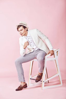 Beyaz ceket, beyaz gömlek, gri pantolon ve açık pembe renkli Kişilik koltuğa oturan beyaz şapka giyen genç yakışıklı mutlu modern meraklı adam 