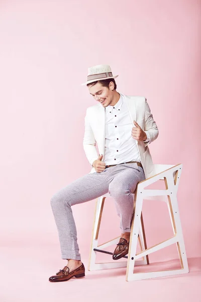 Unga stiliga glada leende moderna människan bär vit jacka, vit skjorta, grå byxor och vit hatt sitter på stolen i studio med ljusrosa bakgrund — Stockfoto