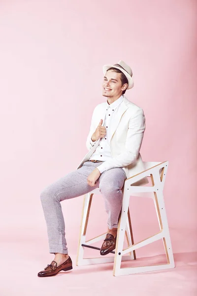 Muda tampan tersenyum tersenyum modern pria mengenakan jaket putih, kemeja putih, celana abu-abu dan topi putih duduk di kursi di studio dengan latar belakang merah muda yang ringan — Stok Foto