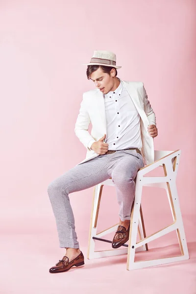 Pemuda tampan bahagia modern pria mengenakan jaket putih, kemeja putih, celana abu-abu dan topi putih duduk di kursi di studio dengan latar belakang merah muda yang ringan — Stok Foto