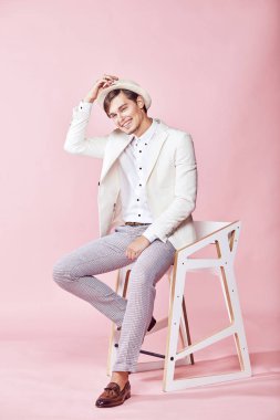 Beyaz ceket, beyaz gömlek, gri pantolon ve açık pembe renkli ile studio sandalyede oturan ve şapka onun elini tutarak beyaz şapka giyen genç yakışıklı mutlu gülümseyen modern insan