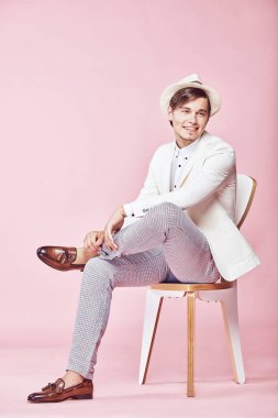 Beyaz ceket, beyaz gömlek, gri pantolon ve açık pembe renkli Kişilik koltuğa oturan beyaz şapka giyen genç güzel mutlu gülümseyen modern erkek modeli 