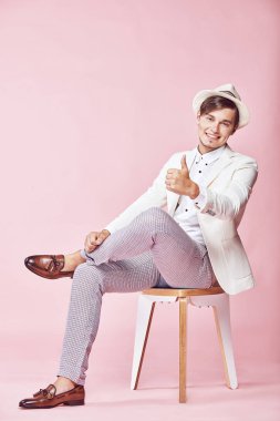 Beyaz ceket, beyaz gömlek, gri pantolon ve açık pembe renkli ile studio sandalyede oturan ve parmağını gösterilmesini beyaz şapka giyen genç yakışıklı mutlu gülümseyen modern insan