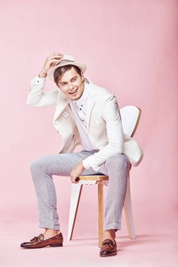 Beyaz ceket, beyaz gömlek, gri pantolon ve açık pembe renkli ile studio sandalyede oturan ve şapka onun elini tutarak beyaz şapka giyen genç yakışıklı mutlu gülümseyen modern insan