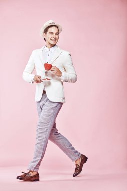 Beyaz ceket, beyaz gömlek, gri pantolon ve açık pembe renkli kişilik aracılığıyla stepping, kırmızı kahve fincanı holding ve uzağa arıyorsunuz beyaz şapka giyen genç güzel mutlu gülümseyen modern erkek modeli