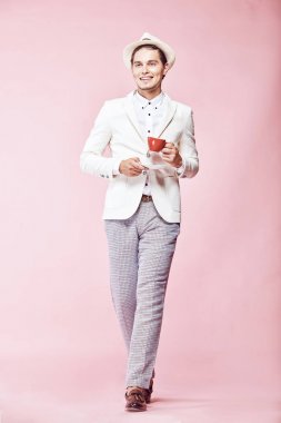 Beyaz ceket, beyaz gömlek, gri pantolon ve açık pembe renkli kişilik aracılığıyla stepping, kırmızı kahve fincanı holding ve uzağa arıyorsunuz beyaz şapka giyen genç güzel mutlu gülümseyen modern insan