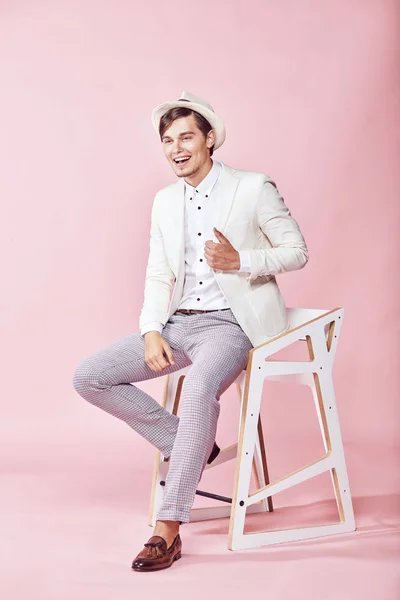 Pemuda tampan modern yang bahagia pria yang bersemangat mengenakan jaket putih, kemeja putih, celana abu-abu dan topi putih duduk di kursi di studio dengan latar belakang merah muda yang ringan — Stok Foto