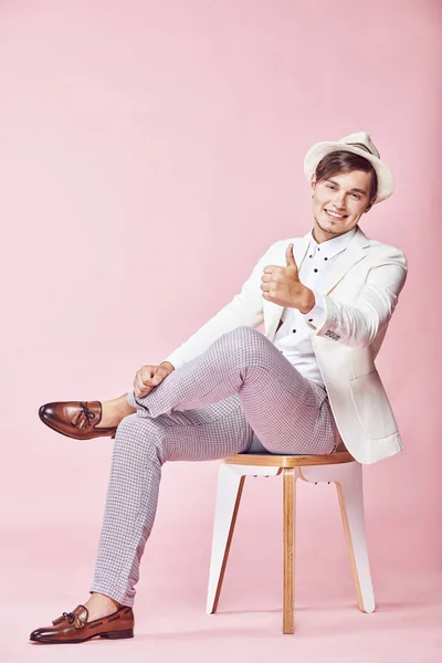Muda tampan tersenyum tersenyum modern pria mengenakan jaket putih, kemeja putih, celana abu-abu dan topi putih duduk di kursi di studio dengan latar belakang merah muda ringan dan menunjukkan jempolnya ke atas — Stok Foto