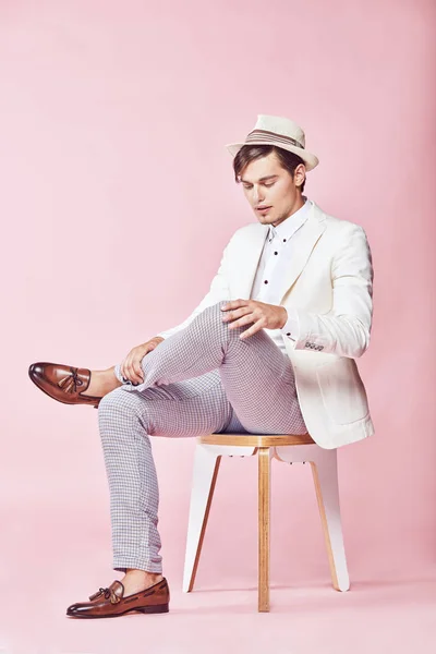 Unga vackra glada leende moderna manlig modell bär vit jacka, vit skjorta, grå byxor och vit hatt sitter på stolen i studio med ljusrosa bakgrund och tittar ner — Stockfoto