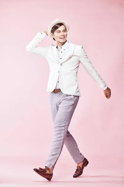 Pemuda cantik tersenyum bahagia modern pria mengenakan jaket putih, kemeja putih, celana abu-abu dan topi putih berpose di studio dengan latar belakang merah muda ringan dan memegang tangannya di topi — Stok Foto