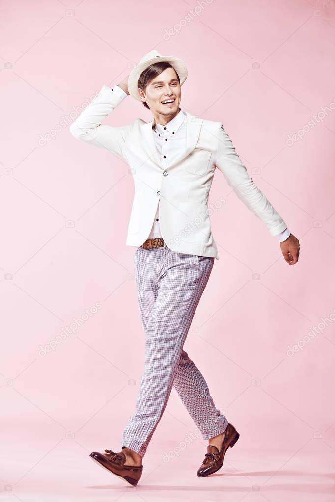 Young beautiful happy smiling modern man wearing white jacket, white shirt, grey pants and white hat posing in studio with light pink background and holding his hand on the hat