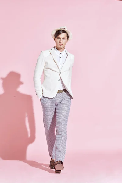 Unga stiliga allvarliga moderna manlig modell bär vit jacka, vit skjorta, grå byxor och vit hatt kliva i studio med ljusrosa bakgrund — Stockfoto