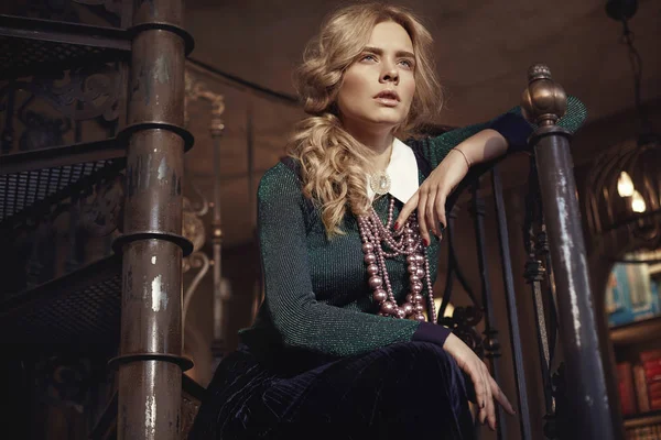 Young beautiful magical blonde curly woman with pearl necklace, wearing green sweater, dark blue skirt, light green tights and shirt with white collar, sitting on dark stairs in fairy room — Stock Photo, Image