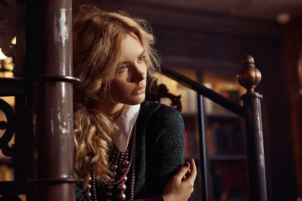 Portrait of young beautiful magical serious blonde curly woman with pearl necklace, wearing green sweater, dark blue skirt, light green tights and shirt with white collar, sitting on dark stairs in fairy room — Stock Photo, Image