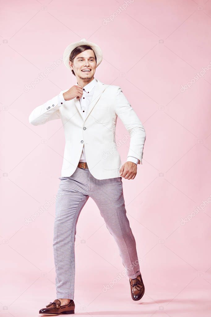 Young beautiful happy smiling modern man wearing white jacket, white shirt, grey pants and white hat stepping in studio with light pink background 