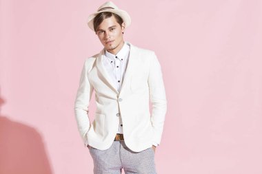 Genç yakışıklı ciddi modern adam beyaz ceket, beyaz gömlek, gri pantolon ve beyaz şapka Studio hafif pembe backgroundc ile poz portresi