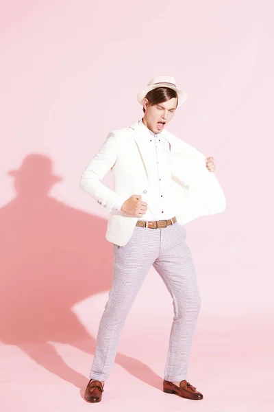 Unga vackra allvarliga moderna människan bär vit jacka, vit skjorta, grå byxor och vit hatt står och poserar i studio med ljusrosa bakgrund — Stockfoto