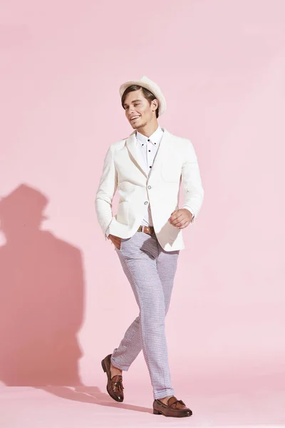 Unga vackra allvarliga moderna människan bär vit jacka, vit skjorta, grå byxor och vit hatt poserar i studio med ljusrosa bakgrund — Stockfoto