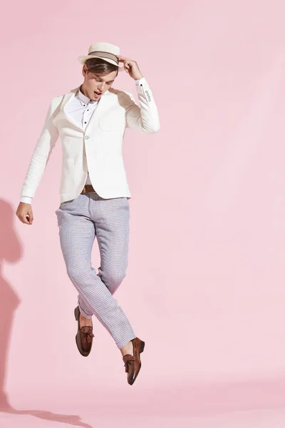 Unga vackra allvarliga moderna människan bär vit jacka, vit skjorta, grå byxor och vit hatt hoppning och poserar i studio med ljusrosa bakgrund — Stockfoto