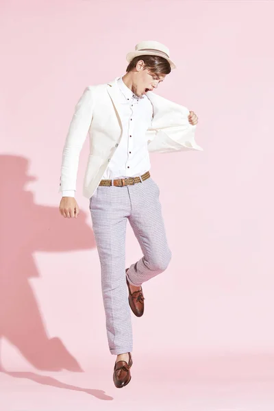 Pemuda tampan serius pria modern mengenakan jaket putih, kemeja putih, celana abu-abu dan topi putih melompat di studio dengan latar belakang merah muda cerah — Stok Foto