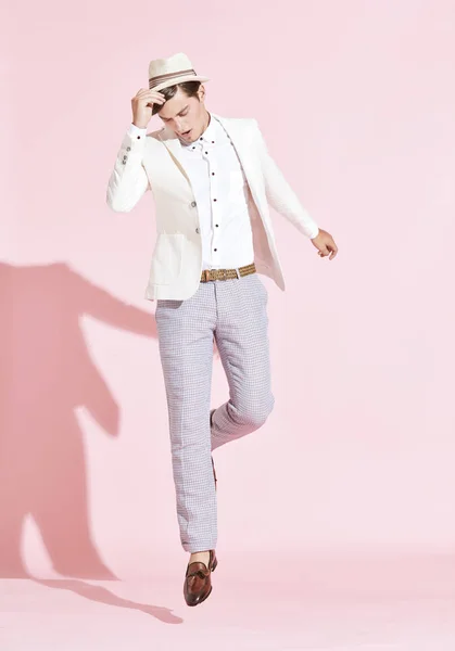 Porträtt av ung stilig allvarliga moderna man bär vit jacka, vit skjorta, grå byxor och vit hatt hoppning i studio med ljusrosa bakgrund — Stockfoto