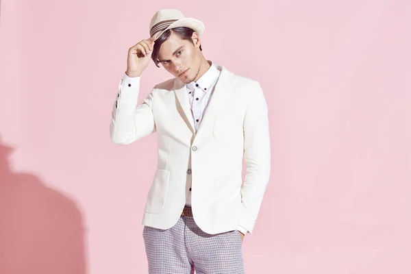 Porträtt av ung stilig allvarliga moderna manlig modell bär vit jacka, vit skjorta, grå byxor och vit hatt poserar i studio med ljusrosa bakgrund — Stockfoto
