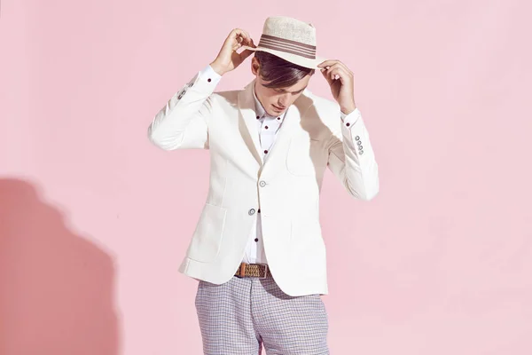 Porträtt av ung stilig allvarliga moderna manlig modell bär vit jacka, vit skjorta, grå byxor och vit hatt poserar i studio med ljusrosa bakgrund — Stockfoto