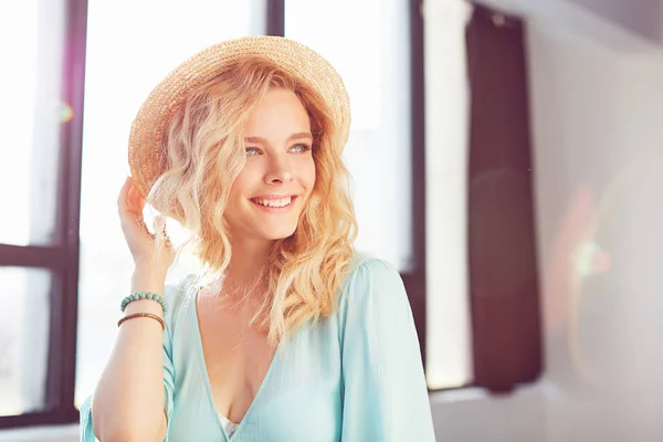 Портрет молодої блондинки сексуальна красива усміхнена жінка з довгим кучерявим волоссям, одягнена в бірюзову сукню, браслети і легкий капелюх позує в сонячній студії в природному світлі — стокове фото