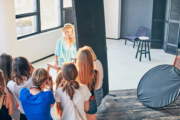 Gli studenti della scuola di fotografia stanno fotografando ritratto di giovane bionda sexy donna incredibile con lunghi capelli ricci, indossando abito turchese, bracciali e cappello leggero in piedi in studio di sole in luce naturale Immagine Stock